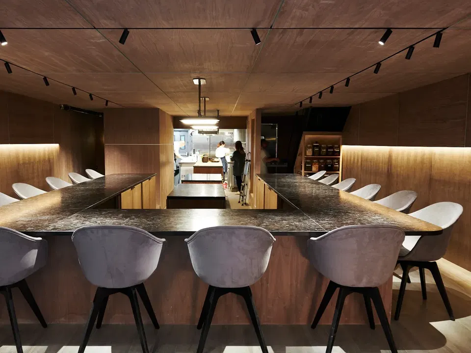 U-shaped dining counter with dark wood top, surrounded by white and gray upholstered chairs with black legs, in a wood-paneled restaurant interior