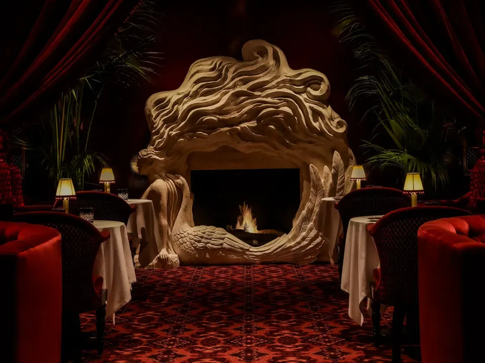 A luxurious lounge with red velvet chairs, decorative carved stone archway with lion motifs, potted plants, table lamps, and red patterned carpeting.