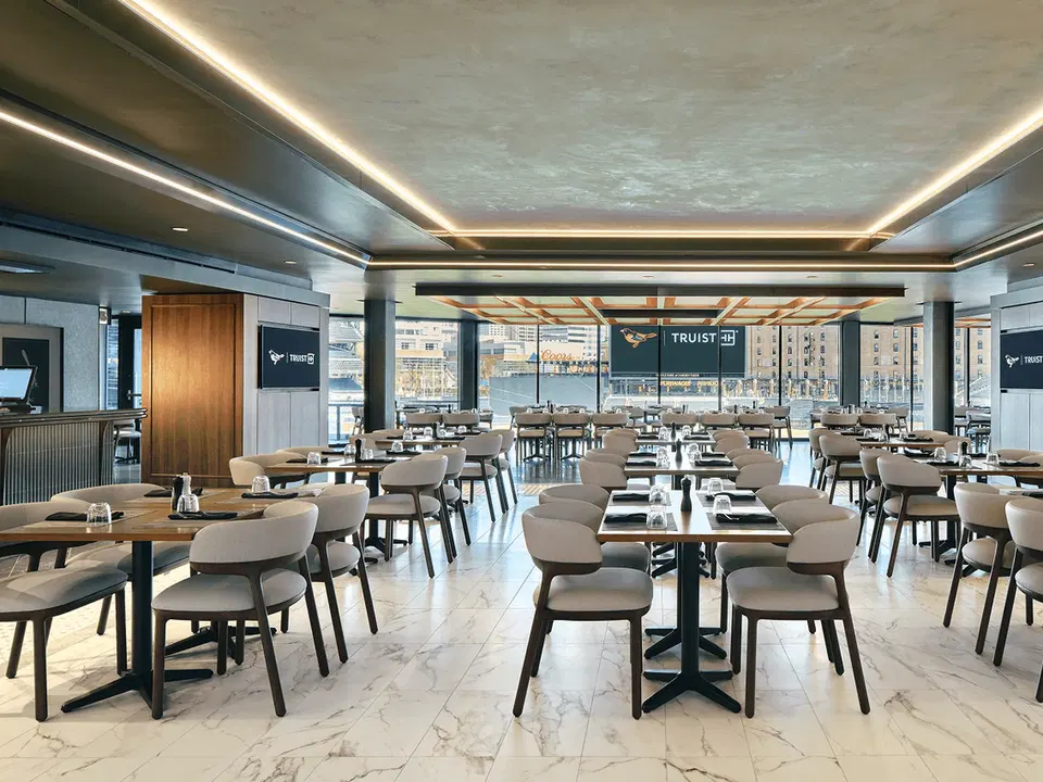 Interior of an upscale restaurant with beige chairs, wooden tables, marble floors, and expansive windows showing an urban waterfront view with buildings in the background.