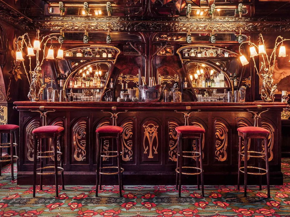An ornate Art Nouveau bar interior with dark mahogany woodwork, gold inlay decorative panels, red velvet bar stools, elaborate branch-form light