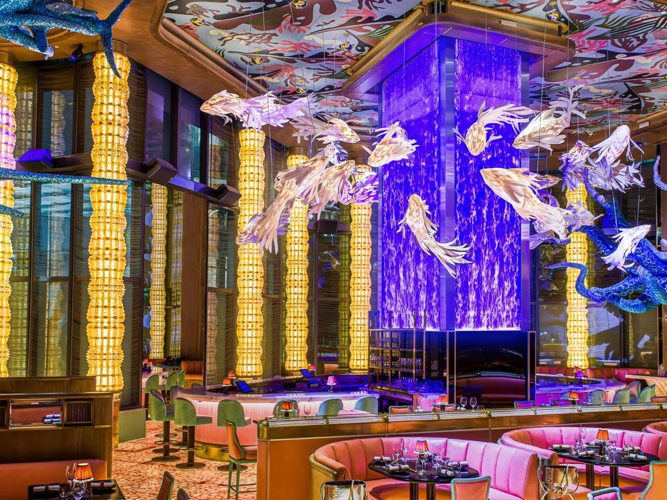 A vibrant restaurant interior with a patterned ceiling, illuminated columns, a purple water feature, and pink banquette seating.