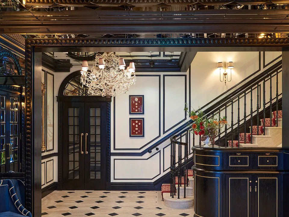 An elegant, dark-toned hotel lobby with a crystal chandelier, black-and-white diamond floor, and a grand staircase with red carpet.