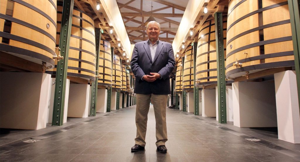A man in a blue blazer and tan pants stands centered in a long corridor lined with large wooden barrels on both sides, with green metal support structures and industrial ceiling.