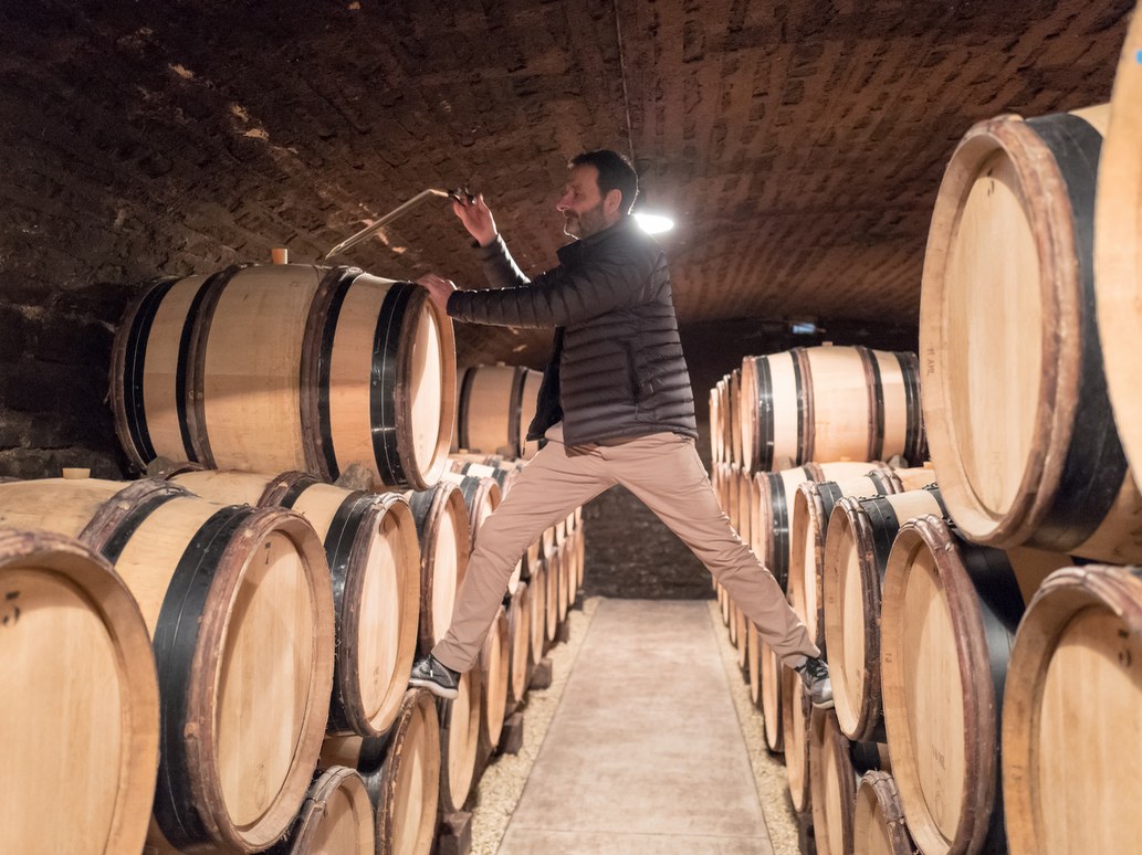 A man in a dark jacket and headlamp stands among wooden wine barrels stacked in rows inside a stone cellar.