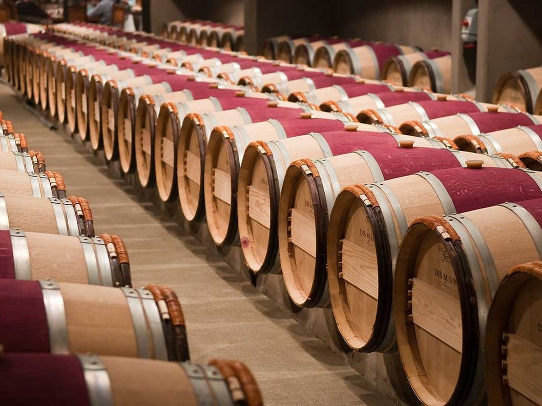 Rows of French oak barrels, many covered in burgundy fabric, are stacked two high in a well-lit winery barrel hall.