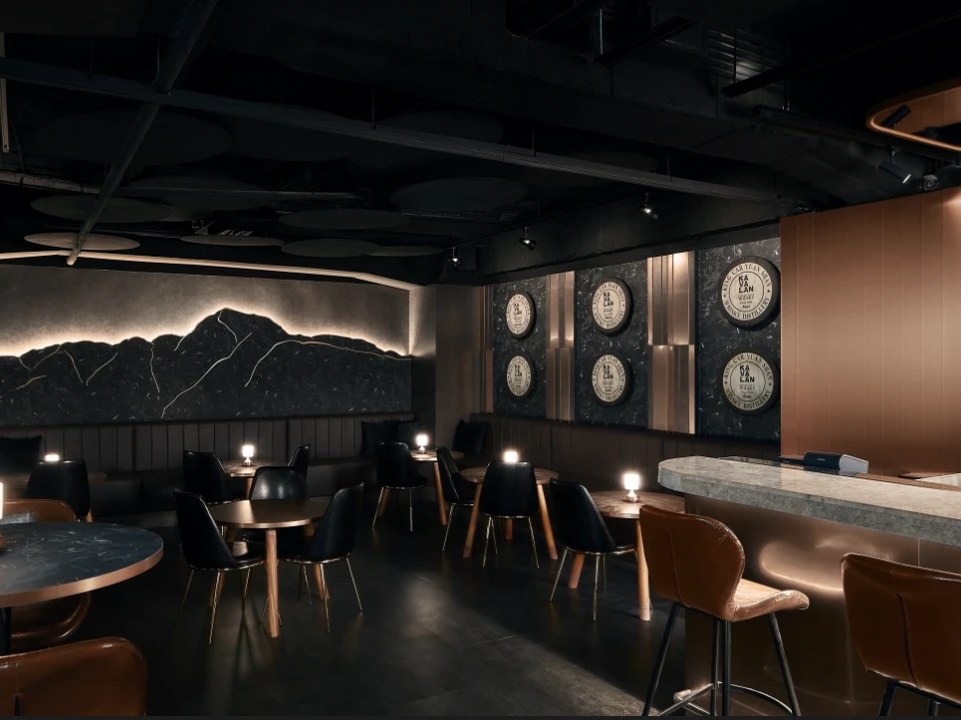A sophisticated dark-toned whisky bar interior with copper accents, barrel medallions on the wall reading 'Ka Lan', a backlit arched bottle display.