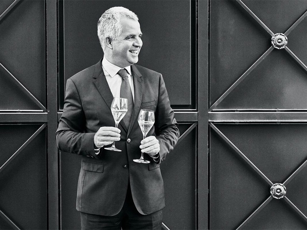 A smiling Olivier Krug, a man with grey hair, holds two champagne flutes filled with sparkling wine, wearing a suit and tie against a dark, geometric