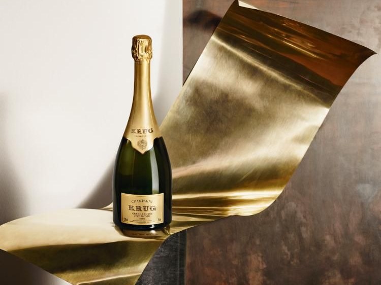 A bottle of Krug Grande Cuvée 173ème Édition Champagne stands on a gold metallic surface against a dark background.