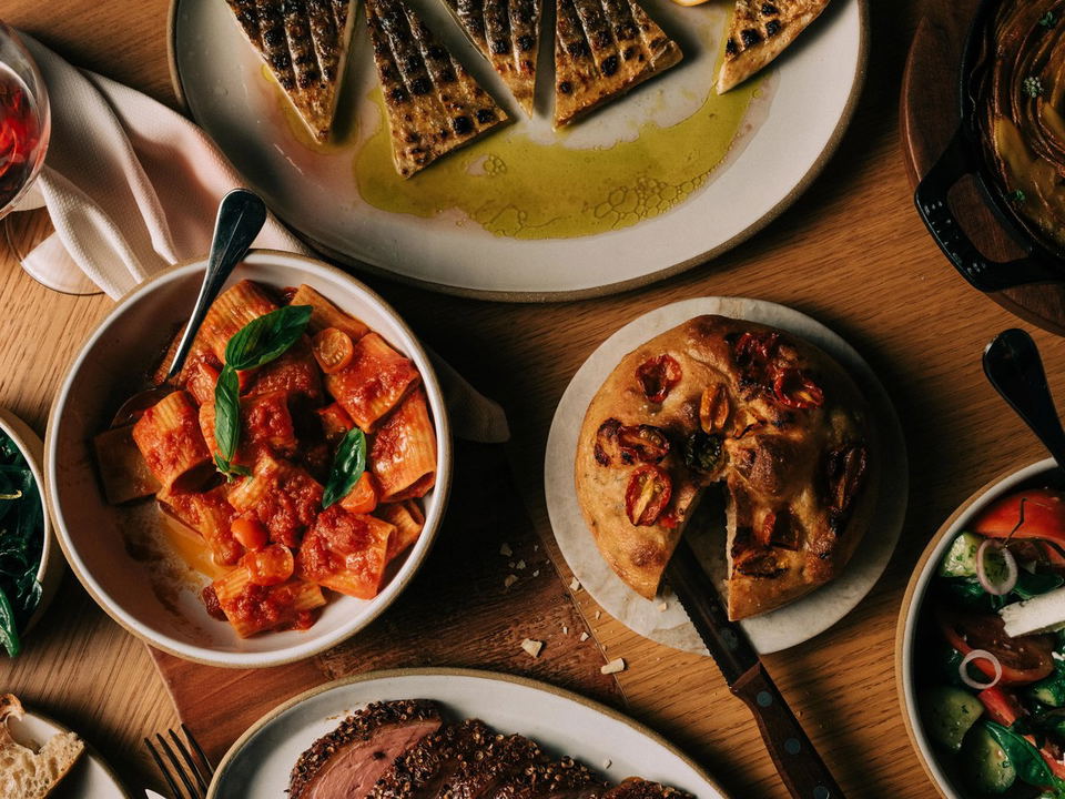 Overhead view of multiple white plates and bowls containing Mediterranean cuisine on a wooden table, including grilled bread, red pepper dishes, and other prepared foods with fresh herbs.