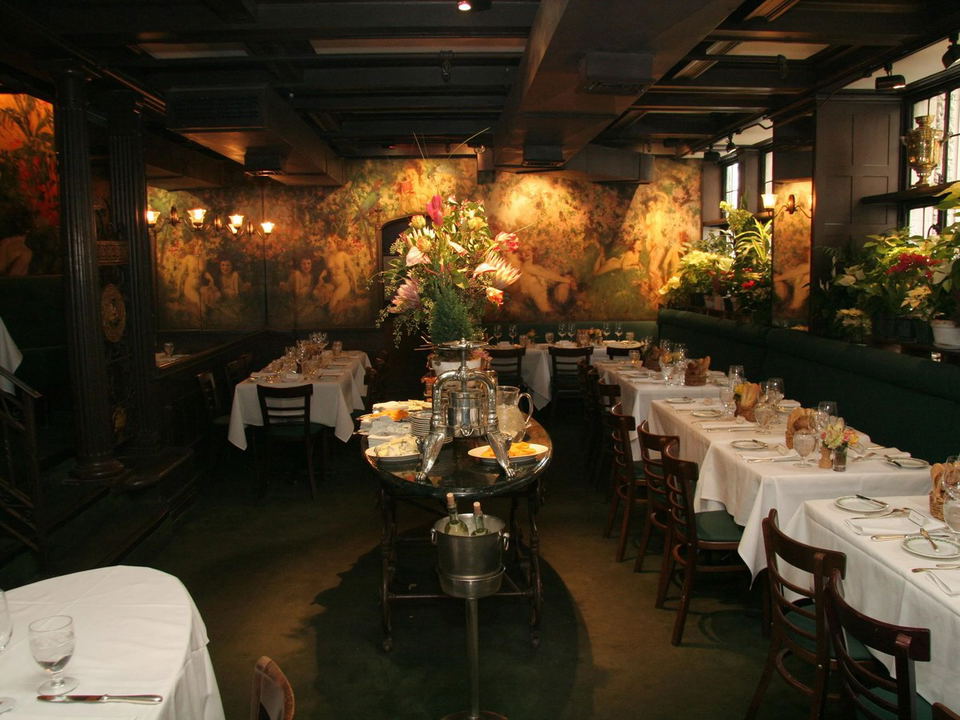 An upscale restaurant dining room with white-tablecloth tables, dark wood chairs, elaborate floral arrangements, and dramatic murals on the walls; the