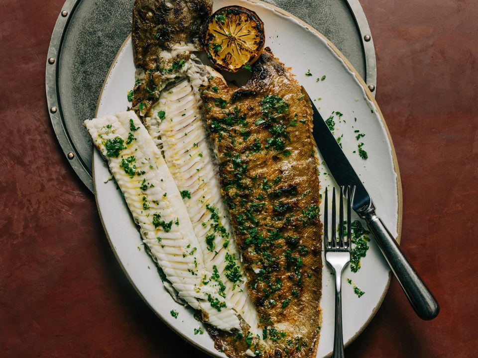 A whole roasted flatfish (likely Dover sole) plated with charred lemon and fresh herbs on an oval white plate with decorative rim, presented on a rust