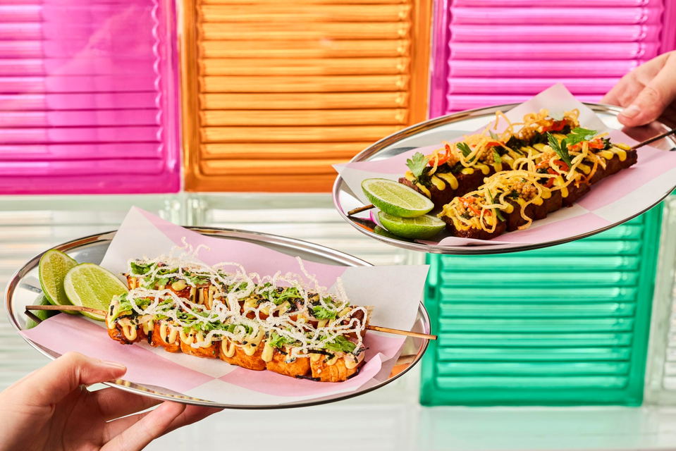 Two hands holding plates of sliced roasted duck skewers garnished with shredded vegetables, fresh cilantro, lime wedges, and yellow crispy noodles, displayed against pink and green neon signage.