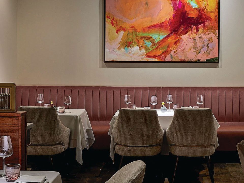 A restaurant interior with a long, plush, dark pink banquette against a light wall, with several tables set with white tablecloths and wine glasses.