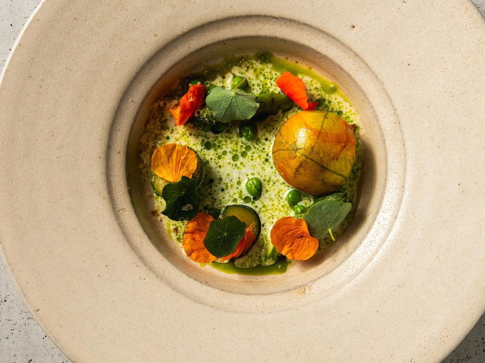 Top-down view of a wide-rimmed cream ceramic bowl containing a green herb emulsion dotted with peas, orange and yellow nasturtium flower petals, green leaves, and a glossy golden sphere at center.