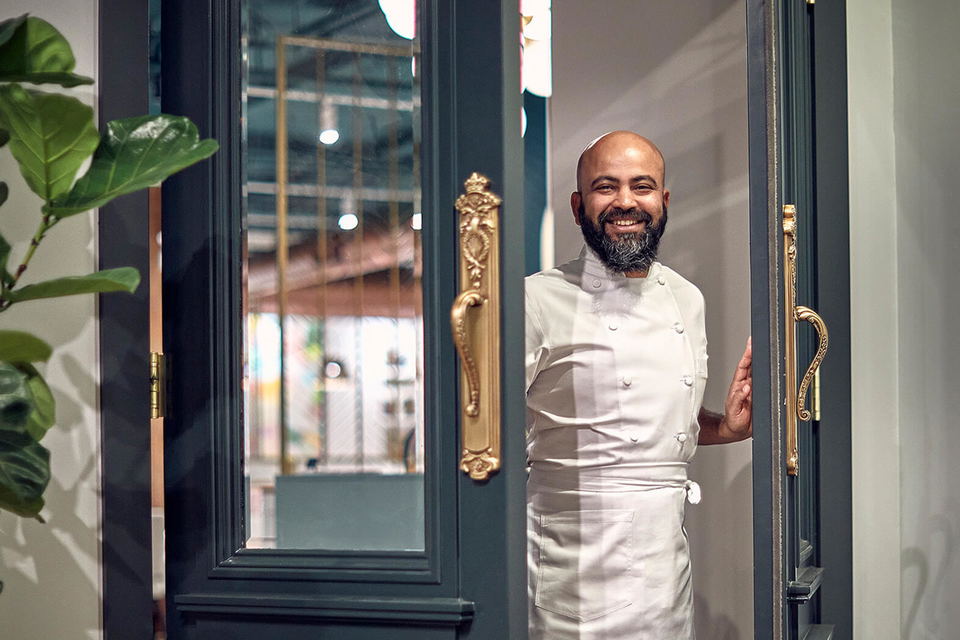 Chef Sujan Sarkar, a bald man with a beard, smiles while standing in a doorway, wearing a white chef's coat and apron.