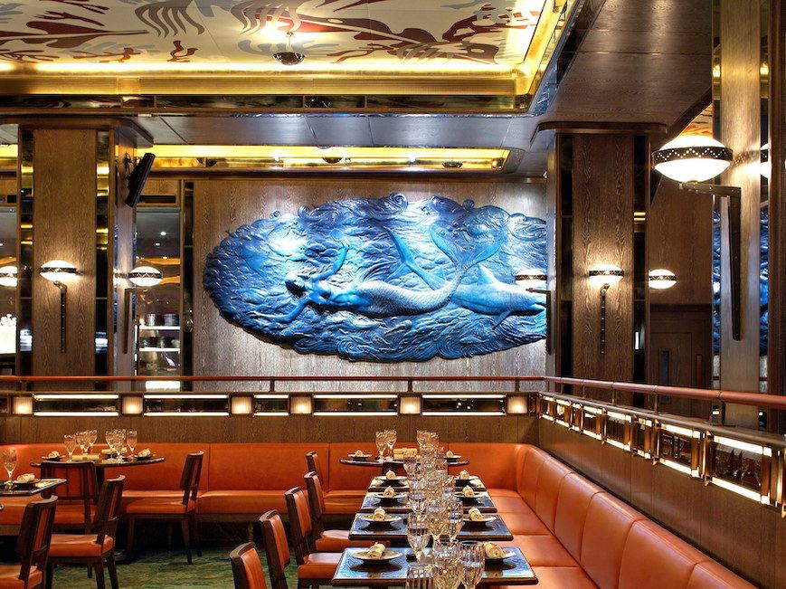 A vibrant blue bas-relief mural of mermaids and sea creatures adorns a wall in Sexy Fish Berkeley Square, with orange banquettes.