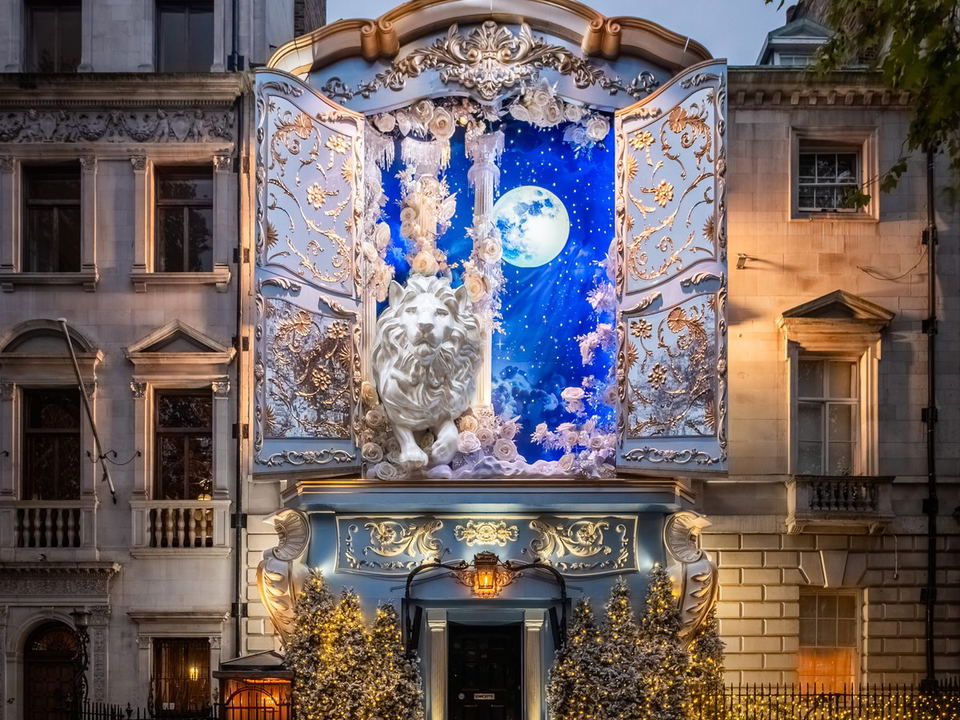 A building exterior at dusk, dramatically decorated with a baroque-style facade, open doors revealing a starry sky, a white lion, and Christmas trees.