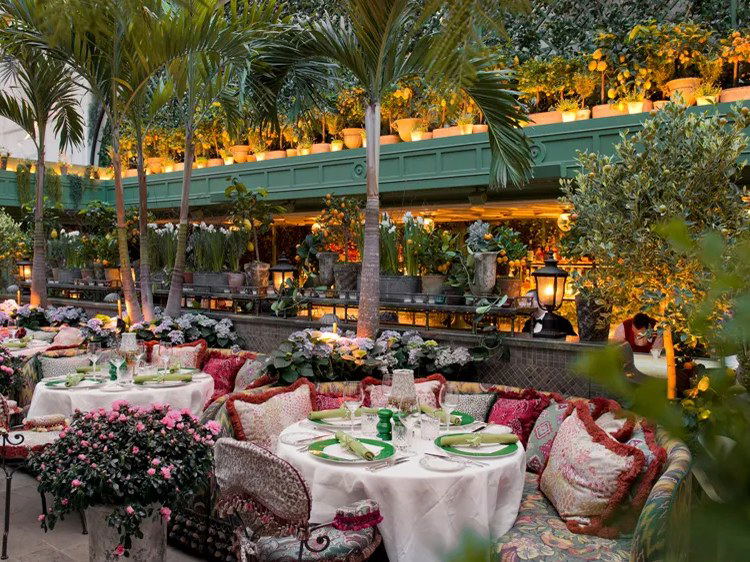 A lush indoor garden terrace with round dining tables, ornate cushioned seating, palm trees, and tiered shelves of potted citrus plants illuminated