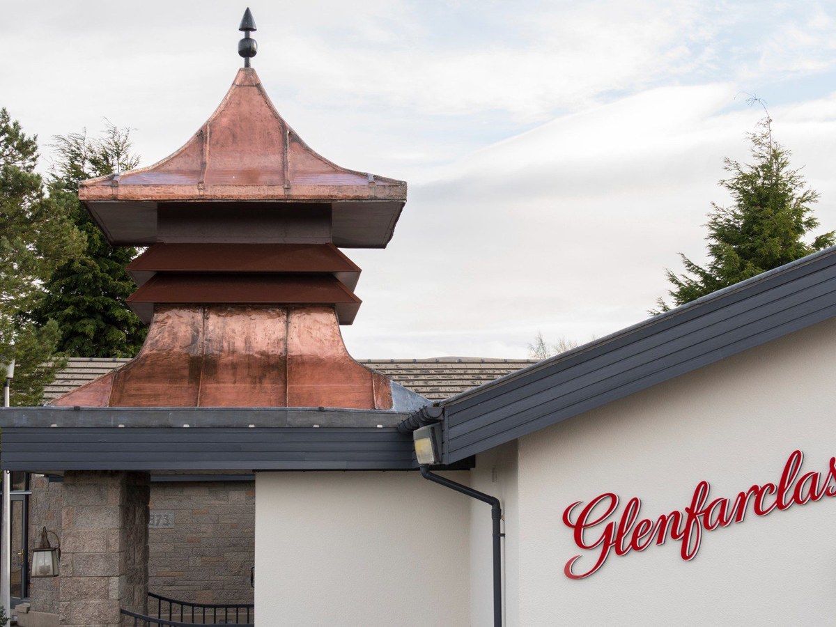 Exterior view of Glenfarclas Distillery's visitor center, featuring a copper pagoda roof and the 'Glenfarclas' name in red script on a white wall.