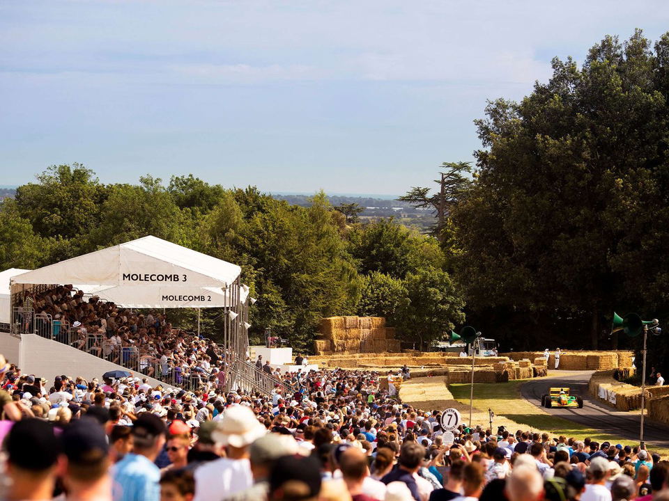 A large crowd of spectators fills the Molecomb 2 and Molecomb 3 grandstands at the Goodwood Festival of Speed, watching a yellow-and-green Benetton.