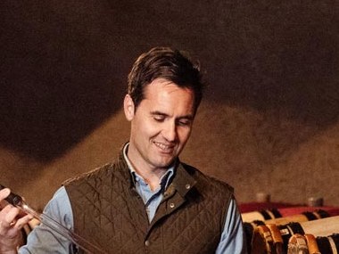 A man in a quilted vest holds a wine thief and a glass of red wine, walking through a barrel cave filled with oak aging barrels.