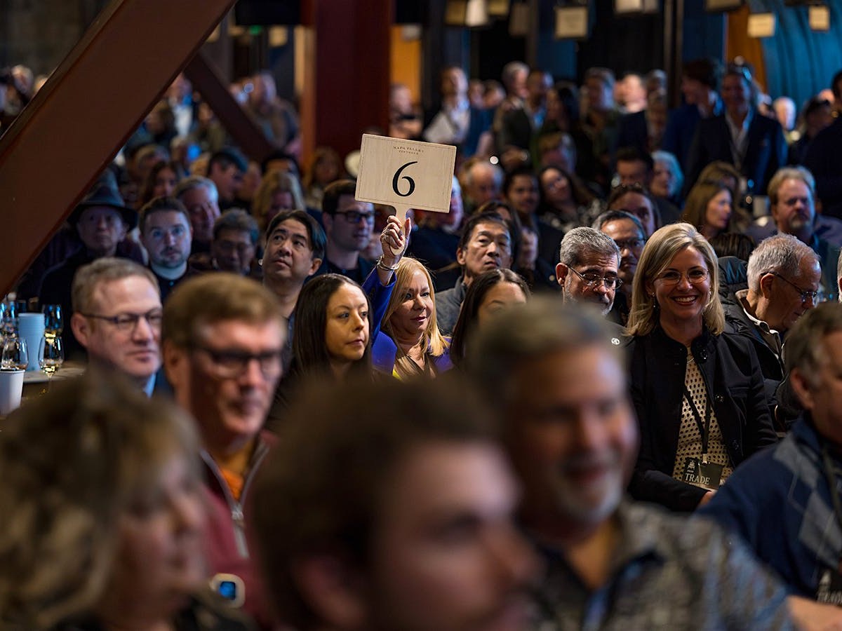 A woman raises a white paddle with the number "6" at a Napa Valley wine auction, surrounded by other attendees.