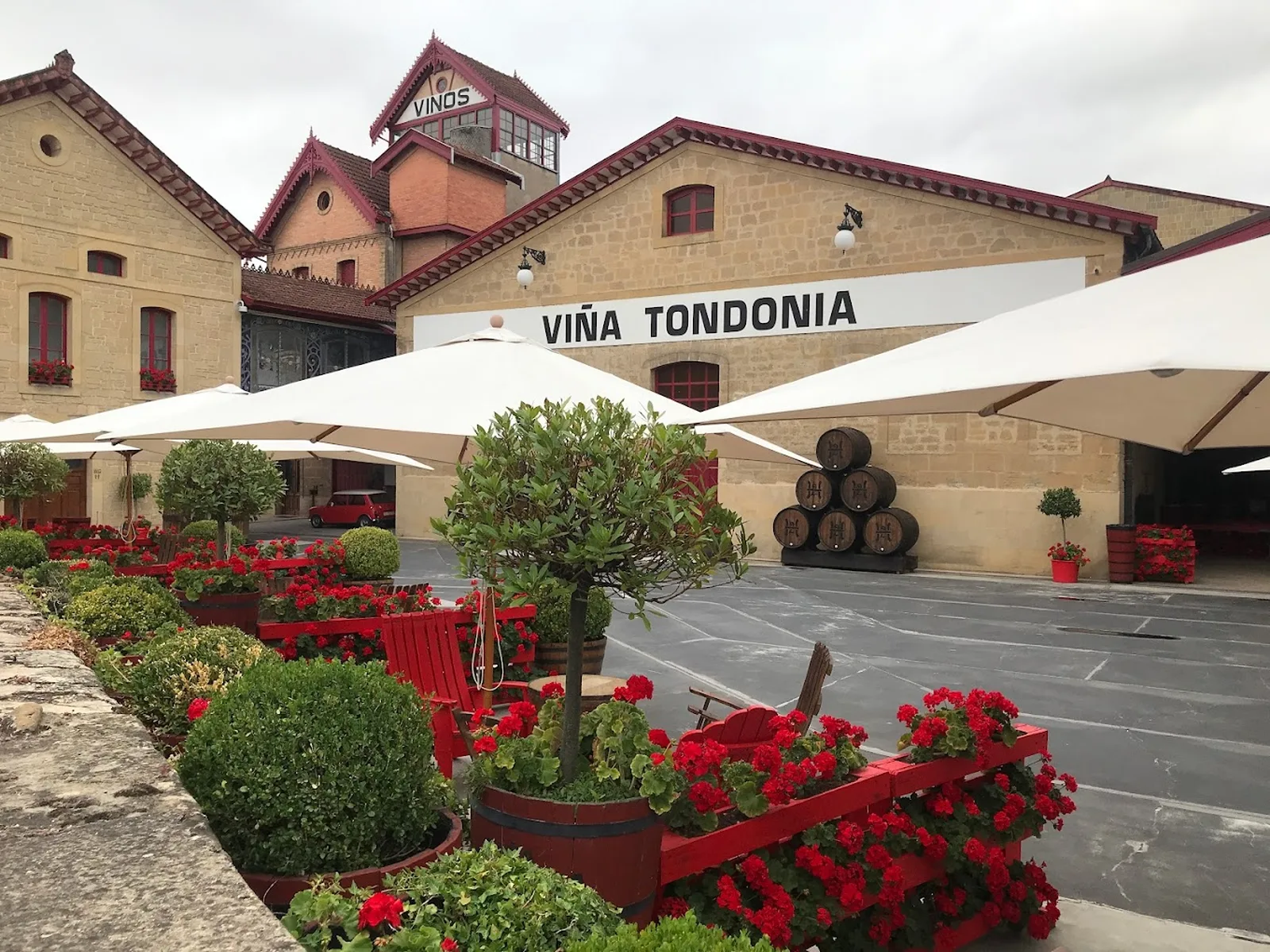 A winery courtyard with beige stone buildings, a tower with VINOS signage, stacked wine barrels, red flowers in planters, white shade umbrellas, and manicured green shrubs.