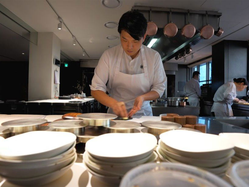 A chef in a white uniform and apron prepares a dish at a counter, surrounded by stacks of white plates in a modern open kitchen.