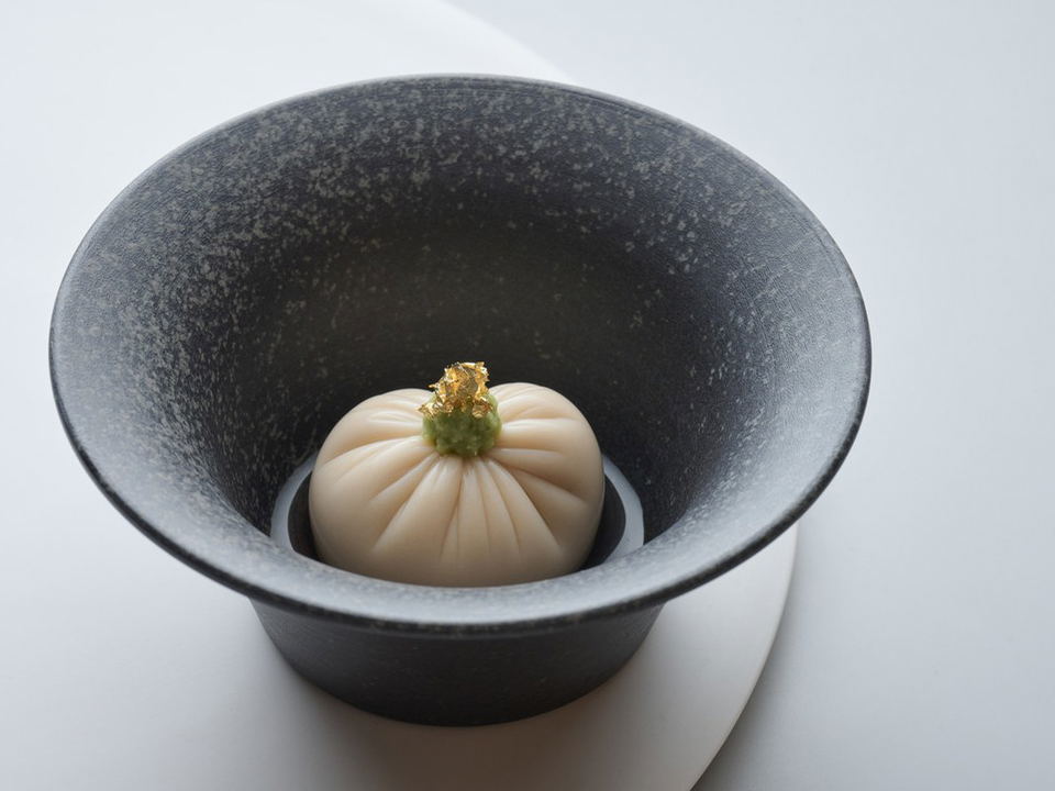 A wagashi-style confection with a ribbed form, green stem, and gold leaf garnish, served in a dark ceramic bowl.