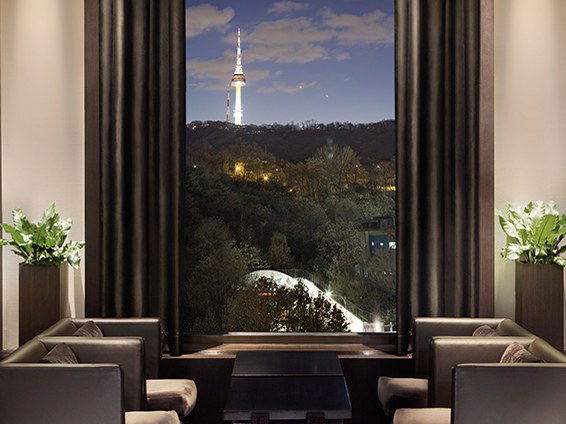 An elegant hotel lounge at The Shilla Seoul at night, with dark curtains, leather seating, potted plants, and a window framing Namsan Tower on a