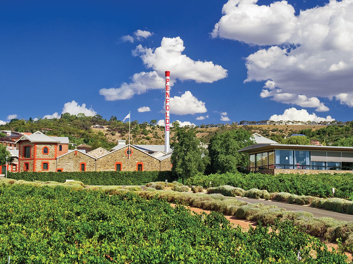 A wide exterior view of Penfolds Magill Estate, showing the iconic white chimney stack with 'PENFOLDS' lettering, historic stone buildings.
