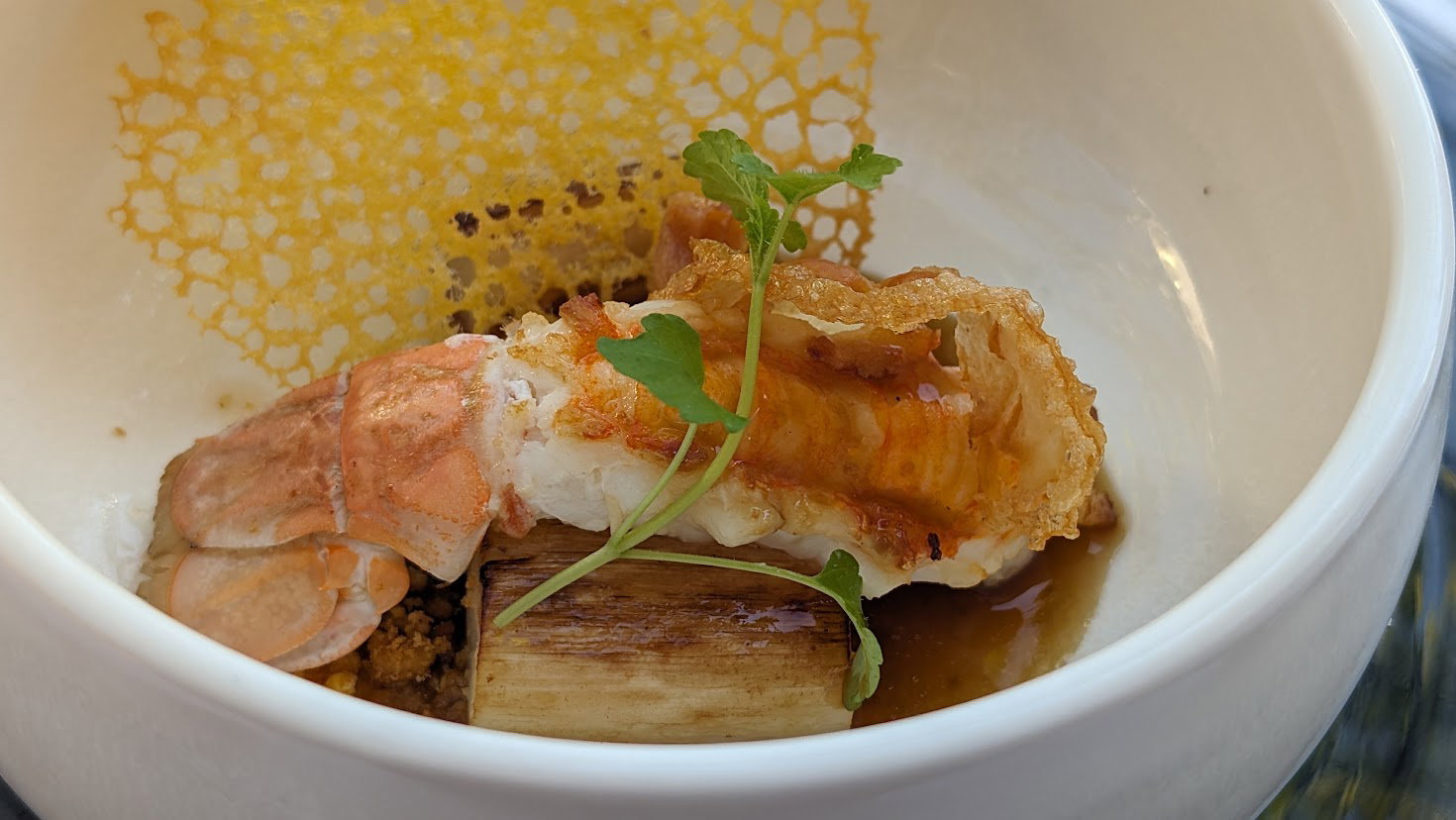 A plated Basque dish featuring what appears to be hake (white fish) with crispy skin, garnished with microgreens and accompanied by a honeycomb tuile,