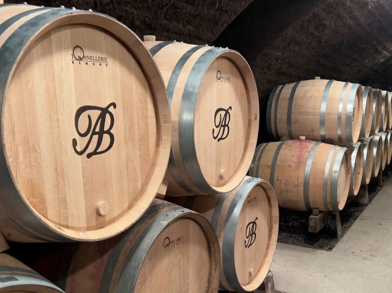Rows of new oak barrels branded with an 'AB' monogram fill a stone wine cellar.