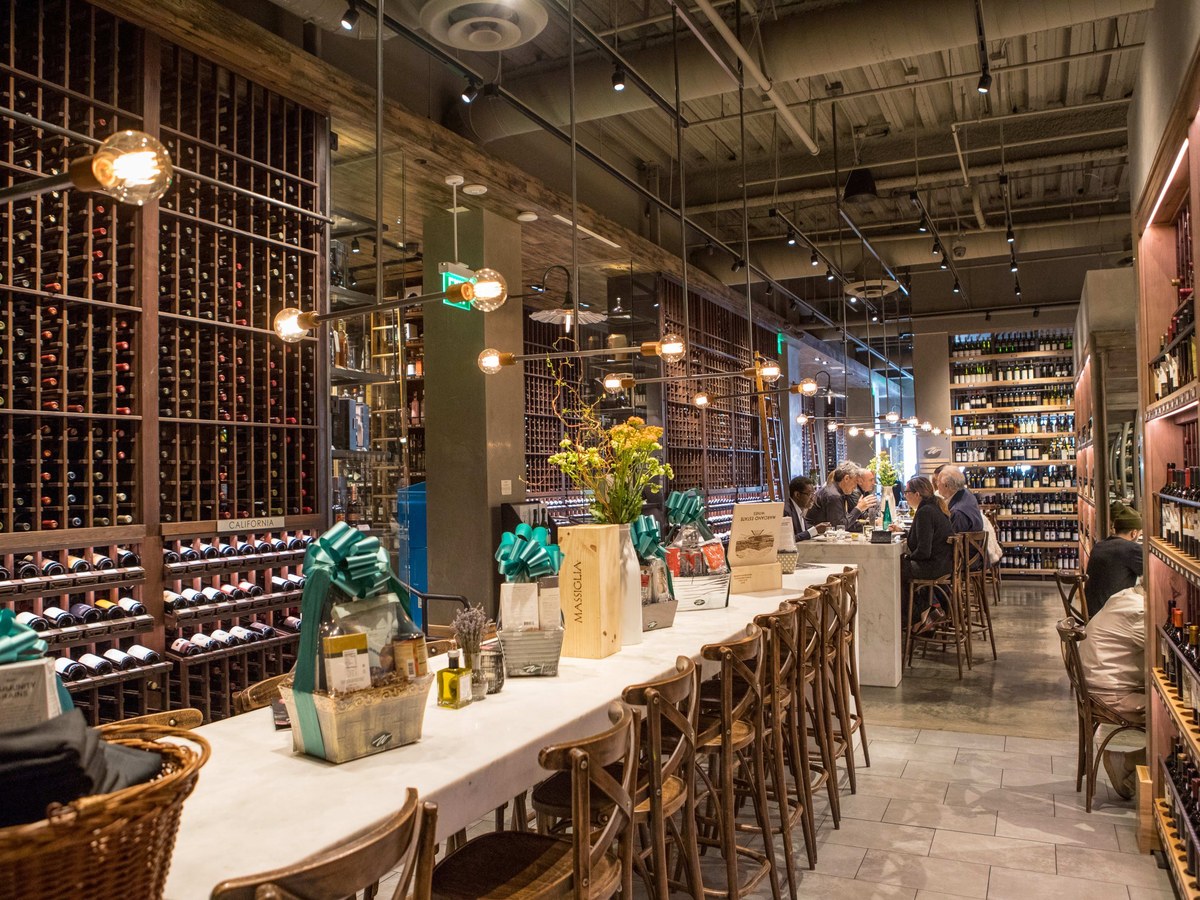Interior of a wine shop and tasting bar with floor-to-ceiling wine racks on both sides, a white counter with wooden chairs, hanging lights, and customers seated at the bar.