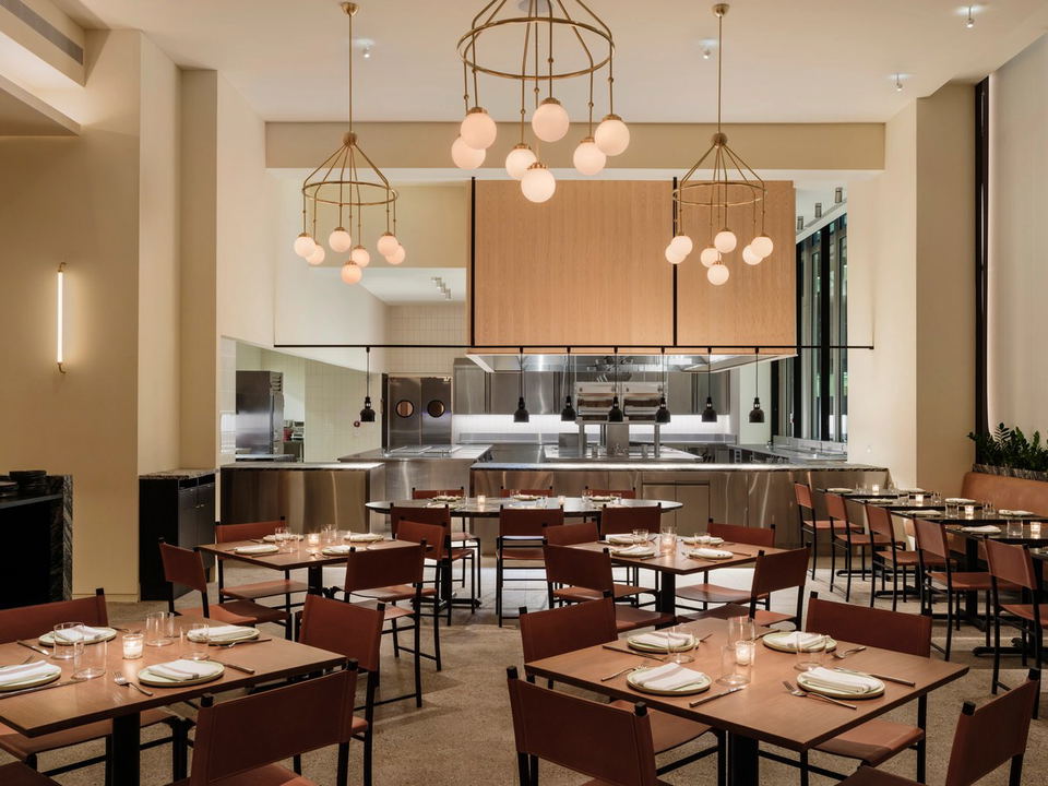 A warmly lit restaurant dining room with an open kitchen, featuring wooden tables set with plates and candles, and chairs upholstered.