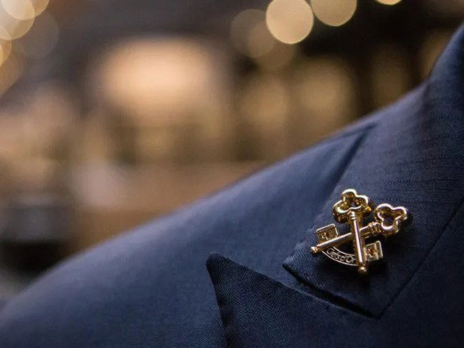 A close-up of a gold Les Clefs d'Or crossed keys pin on the lapel of a navy herringbone suit.