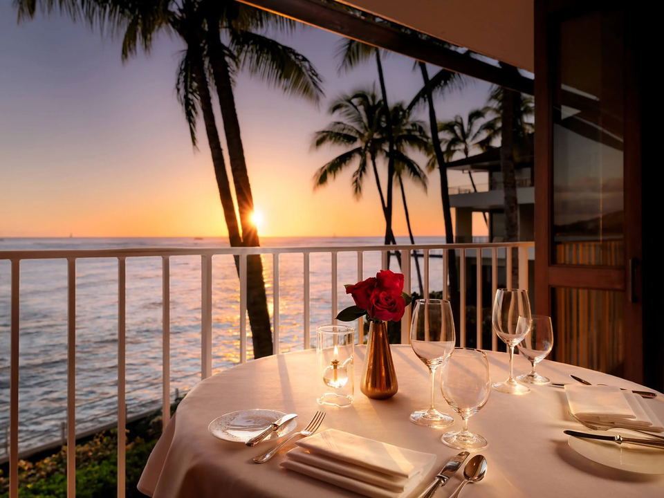 An elegantly set dining table for two on an oceanfront terrace at sunset, with palm tree silhouettes, candlelight, a red rose centerpiece, and wine.