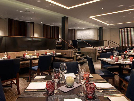 An elegantly set fine-dining room interior with navy upholstered chairs, dark wood tables, crystal wine glasses, red cut-glass tumblers, and warm.