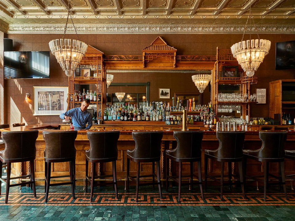A bartender in a plaid shirt pours a drink at the J-Bar, a historic bar with a pressed-tin ceiling, crystal chandeliers, and leather bar stools.