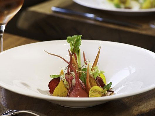 A beautifully plated fine-dining dish featuring roasted heirloom beets and baby carrots with microgreens in a wide white bowl, served in an upscale.