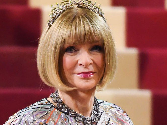 Anna Wintour, with a blonde bob and a jeweled tiara, wears an elaborate feathered cape gown while standing on red-carpeted stairs.
