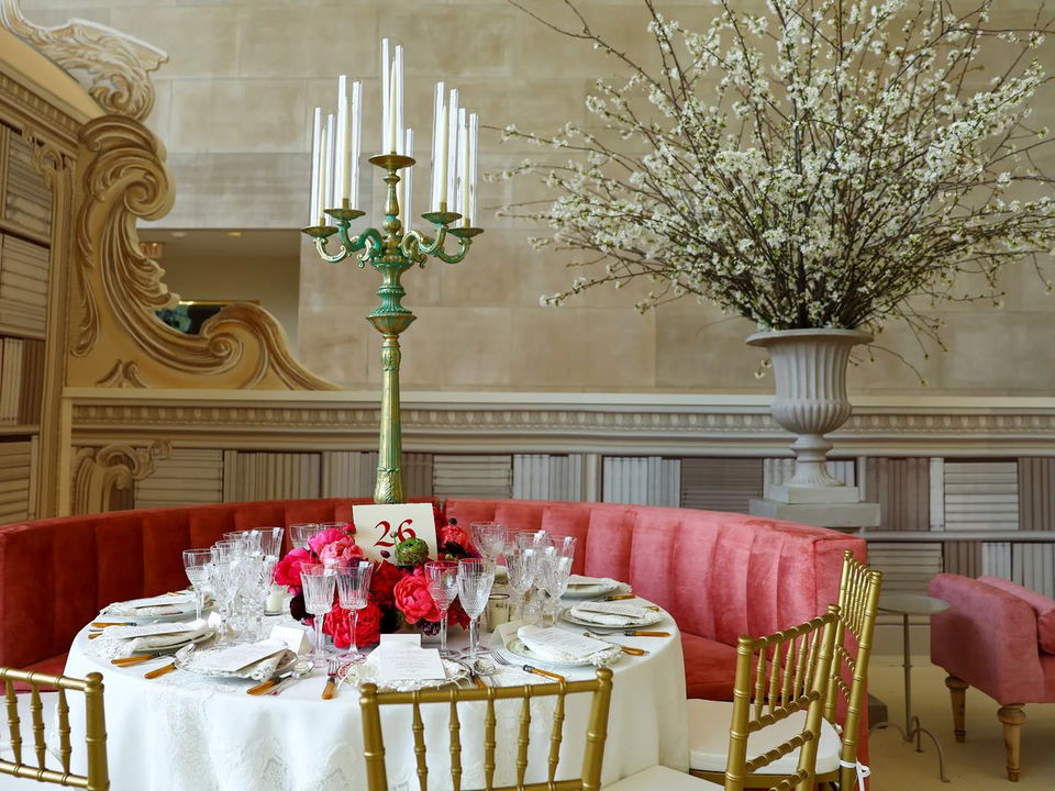 An elegantly set round dinner table with crystal glassware, pink floral centerpiece, tall green candelabra, and gold chiavari chairs inside a grand