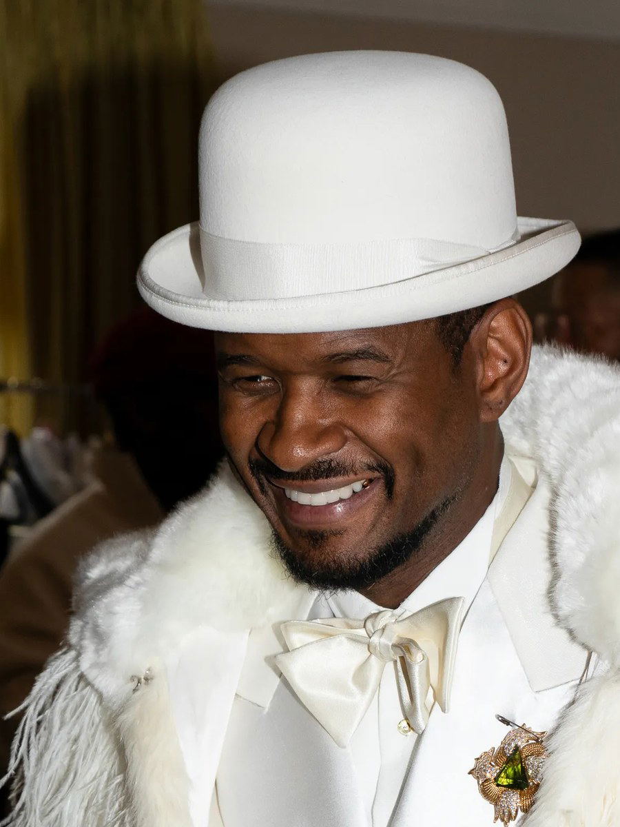 Usher, dressed in an all-white ensemble including a bowler hat, feathered coat, and suit, smiles while holding a gold-handled cane.