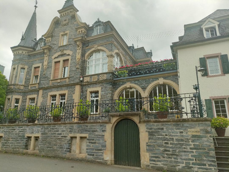 A multi-story stone building with green shutters, mansard roofs, and decorative ironwork balconies, situated on a hillside with manicured grounds.
