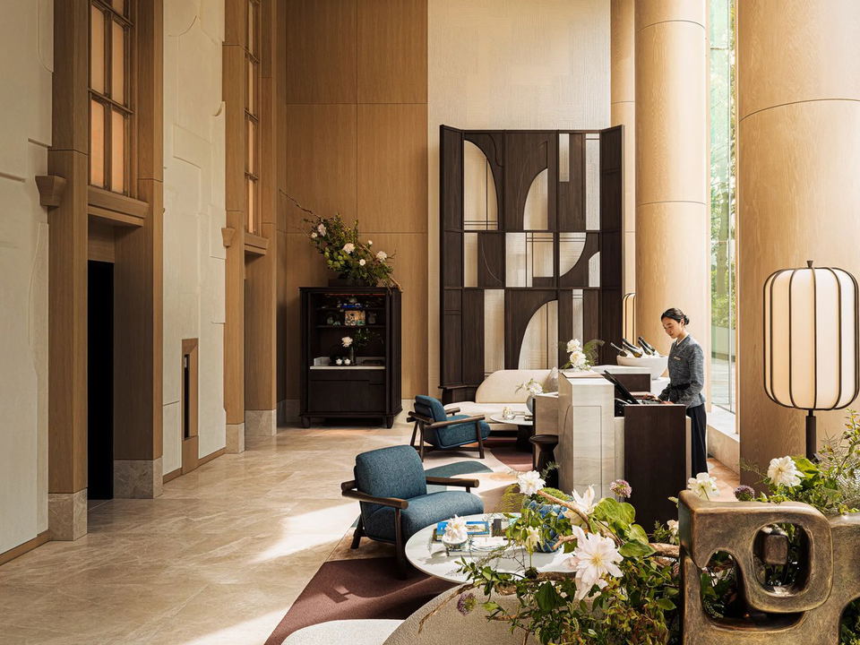 A hotel lobby with a staff member at a reception desk, blue armchairs, a decorative screen, tall columns, and floral arrangements.