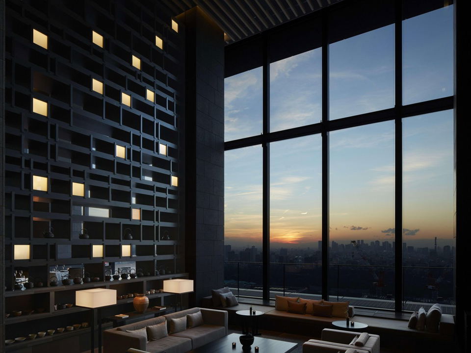 A modern, dimly lit lounge with floor-to-ceiling windows overlooking a city skyline at sunset. An illuminated grid wall features small, glowing