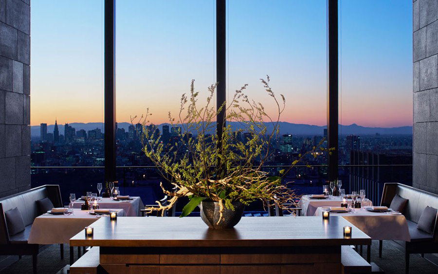 An upscale restaurant dining room at twilight with floor-to-ceiling windows overlooking a Tokyo cityscape, featuring a dramatic floral arrangement.