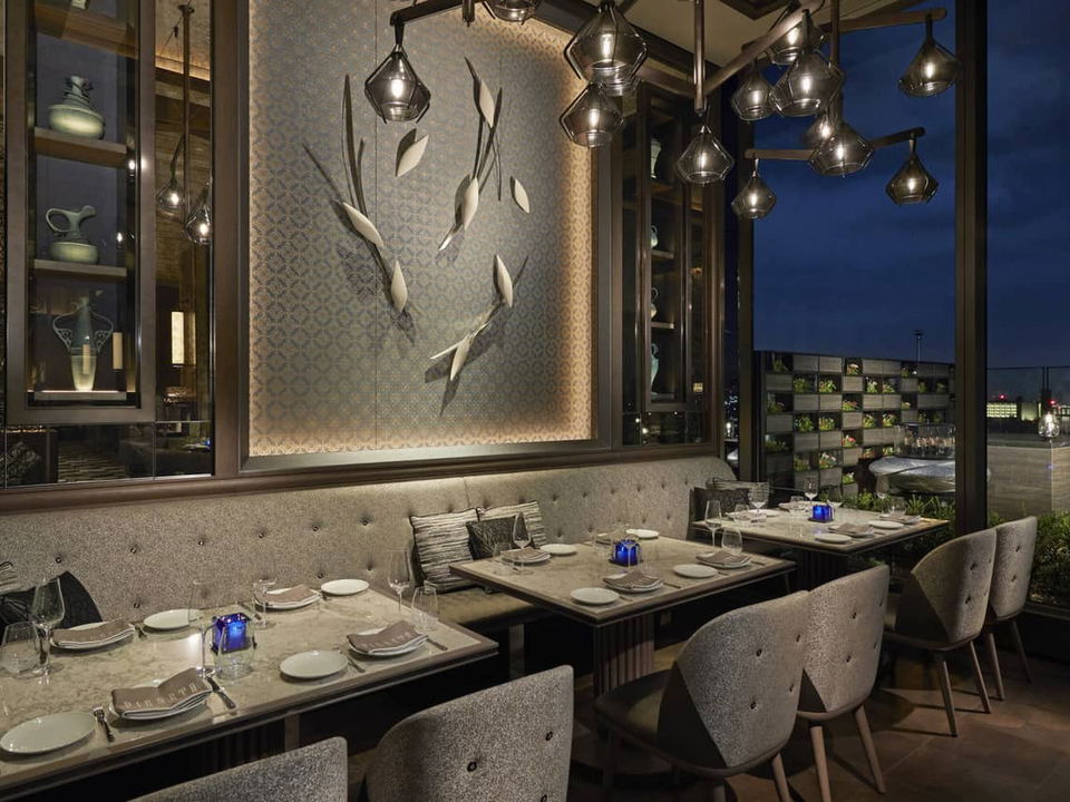 An elegant restaurant dining room at night with tufted banquette seating, geometric pendant lights, decorative wall art, and a city skyline.