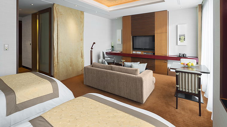 A luxury hotel suite interior with two beds, a living area with sofa and entertainment center, and modern furnishings.