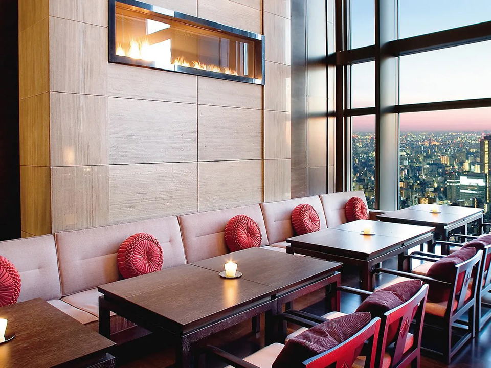 An elegant high-rise restaurant dining room with low dark-wood tables, cream banquette seating with red round cushions, a wall-mounted linear
